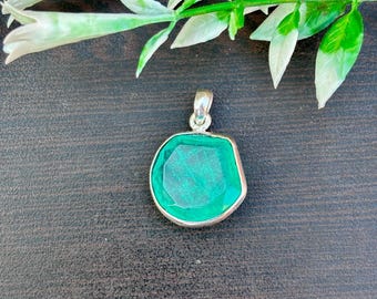 May include: A silver pendant featuring a large, faceted emerald-green gemstone. The pendant has a simple design, with the gemstone set in a silver bezel. The background is a dark, textured surface with green and white leaves.