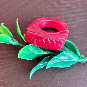 Op de afbeelding: Een gesneden, robijnrode ring met een geometrisch ontwerp. De ring wordt getoond op een bed van heldergroene, kunstmatige bladeren. Het oppervlak van de ring heeft een gestructureerd, geribbeld patroon en de kleur is een levendig, doorschijnend rood.