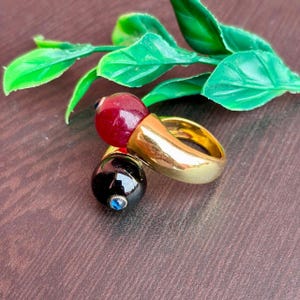 Puede incluir: Dos anillos con bandas doradas. Uno presenta una cuenta roja oscura y el otro una cuenta negra con un acento azul. Los anillos se exhiben sobre una superficie marrón con hojas verdes en el fondo.