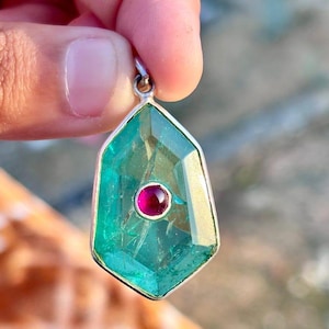 May include: A faceted, shield-shaped pendant featuring a translucent teal-green gemstone set in a silver frame. A small, circular, deep pink gemstone is bezel-set in the center of the larger teal stone, which hangs from a silver bail.
