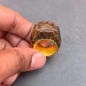 以下が含まれることがあります： 厚みのあるファセットカットされた茶色と黒の石のトップが付いたリングで、表面はざらざらとした質感です。リングの内側は滑らかな明るいイエローゴールド調の金属バンドで裏打ちされており、縁には装飾的なスカラップエッジが施されています。