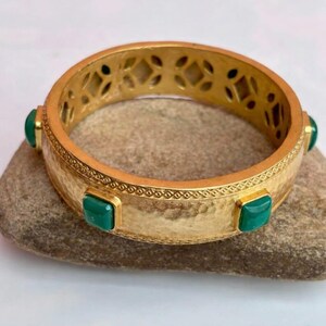 Könnte beinhalten: Ein goldfarbenes Armreif-Armband mit gehämmerten Texturen und geometrischen Ausschnitten. Das Armband hat quadratische, smaragdgrüne Edelsteine. Das Armband liegt auf einer Steinoberfläche.
