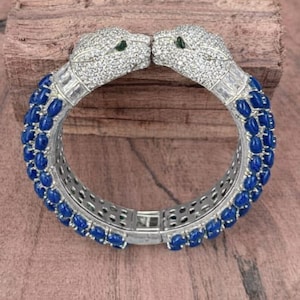 May include: A silver-toned bracelet featuring two panther heads encrusted with clear crystals and green eyes. The bracelet band is adorned with blue gemstones. The jewelry is set against a wooden surface.