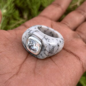 May include: A chunky ring with a white and gray speckled stone. The ring features a square, clear gemstone set in a silver bezel. The ring is held in a hand.