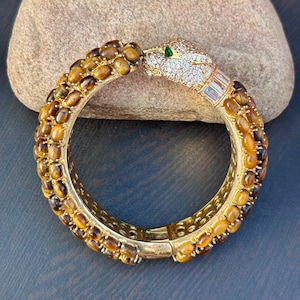 Puede incluir: Brazalete dorado con forma de serpiente con piedras ovaladas marrones y amarillas. La cabeza de la serpiente está incrustada con cristales transparentes y un ojo verde esmeralda. El brazalete es articulado.