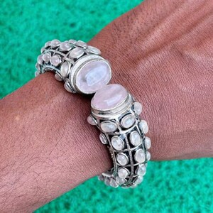 以下が含まれることがあります： ヒンジデザインのシルバーブレスレット。大きな楕円形のローズクォーツと、小さく丸い透明な石が特徴です。 ブレスレットは、緑色の背景で、人の手首に表示されています。