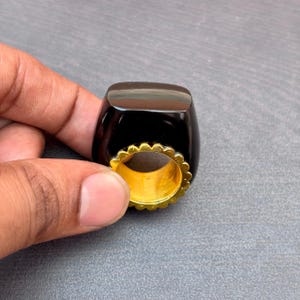 以下が含まれることがあります： 大きな滑らかで磨かれた黒い石がゴールドトーンの金属バンドにはめ込まれた指輪。バンドには指を通す開口部を囲む装飾的なスカラップ状の縁があり、暗い石とゴールドのコントラストを生み出しています。