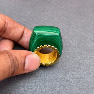以下が含まれることがあります： 鮮やかな濃い緑色の縞模様が入った、滑らかで長方形の緑色のマラカイト石をあしらった指輪。石は、石座の周囲に装飾的なスカラップ状の縁取りが施されたゴールドトーンの金属製バンドにはめ込まれています。