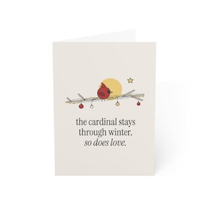 May include: A greeting card with a cream background featuring a red cardinal perched on a pine branch decorated with small ornaments, a yellow sun, and a star. The text reads: the cardinal stays through winter. so does love.