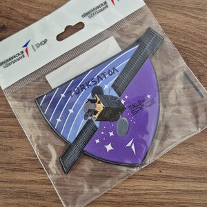 May include: A patch featuring a satellite design with the text "TURKSAT 6A" and "TALES of SPACE" in a plastic sleeve. A white golf ball and a decorative white golf ball are also in the image.