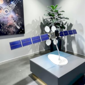 Könnte beinhalten: Ein Modell eines Satelliten mit blauen Sonnenkollektoren und weißen, kreisförmigen Antennen. Der Satellit steht auf einem Ständer auf einer grauen Plattform. Ein Gemälde einer Raumstation ist zu sehen.