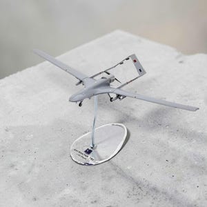 May include: A grey Bayraktar TB2 unmanned aerial vehicle model mounted on a clear stand. The base features the text "BAYRAKTAR TB2 1/32 Ölçek". The aircraft has a long wingspan, a rear propeller, and twin vertical stabilizers with a small flag emblem.