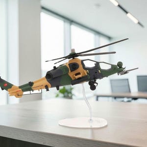 May include: A detailed model helicopter in tan, green, and black camouflage colors, mounted on a clear stand. The model features a rotor, tail, and intricate detailing, suggesting a military or aviation theme. The helicopter is displayed on a light-colored table.