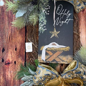 May include: A Christmas wreath with a wooden base, adorned with greenery, gold and blue ribbons, and a sign that reads "O Holy Night." The sign features a gold star and a nativity scene.