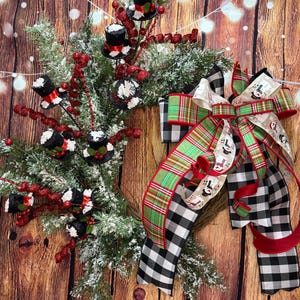 May include: A festive Christmas wreath with faux snow-covered pine branches. It features black top hat ornaments with red accents, red berry sprigs, and a large bow with plaid and snowman-themed ribbons. The background is a wooden surface.