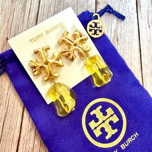 May include: Gold-tone earrings with a signature logo design and yellow, rectangular, dangling accents. The earrings are displayed on a white card with the text "TORY BURCH" and a blue drawstring bag with the brand's logo.