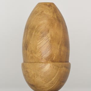 May include: A smooth, light brown wooden object with a distinct egg-like shape and a visible horizontal seam dividing the top and bottom sections. The wood grain displays swirling patterns in varying shades of tan and golden brown throughout the surface.