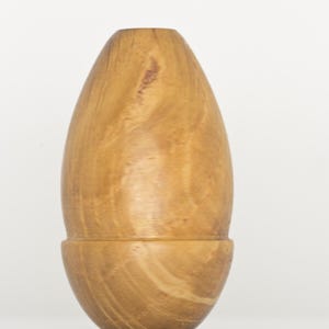 May include: A smooth, light brown wooden vessel with a rounded, egg-like shape. The object features a horizontal seam separating a larger upper section from a smaller, rounded base, showcasing natural wood grain patterns throughout the surface.
