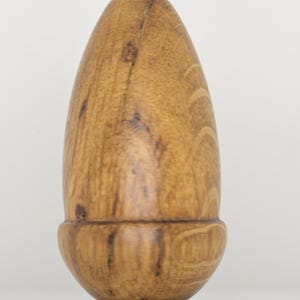 May include: A smooth, light brown wooden object shaped like an acorn, featuring a rounded base and a tapered top. The wood grain is visible throughout the surface, displaying natural variations in colour and texture with a polished finish.