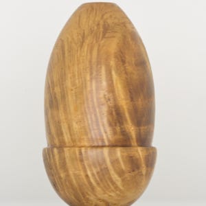 May include: A smooth, light brown wooden object shaped like an acorn with a rounded base and a tapered top. The surface displays a natural wood grain pattern with darker brown streaks running vertically throughout the entire piece.