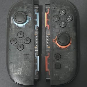 Puede incluir: Dos mandos de juego negros transparentes, que revelan componentes internos. El mando izquierdo tiene un contorno de joystick azul, el derecho naranja. Cada uno tiene botones negros y pads direccionales. Accesorio de juego moderno.