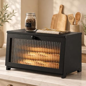 May include: A black wooden bread box featuring a rectangular frame and a front-facing door with vertical ribbed glass panels. The box has a small round knob for opening and is positioned on a countertop, showcasing a clear view of the interior contents.