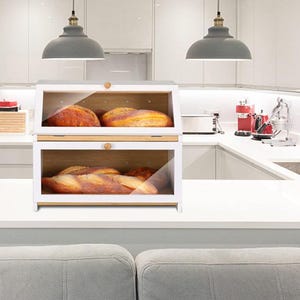 May include: A two-tiered white bread box with a clear glass front and a wooden interior. The box is filled with loaves of bread. The top of the box has a wooden handle. The bread box is on a white countertop in a kitchen.