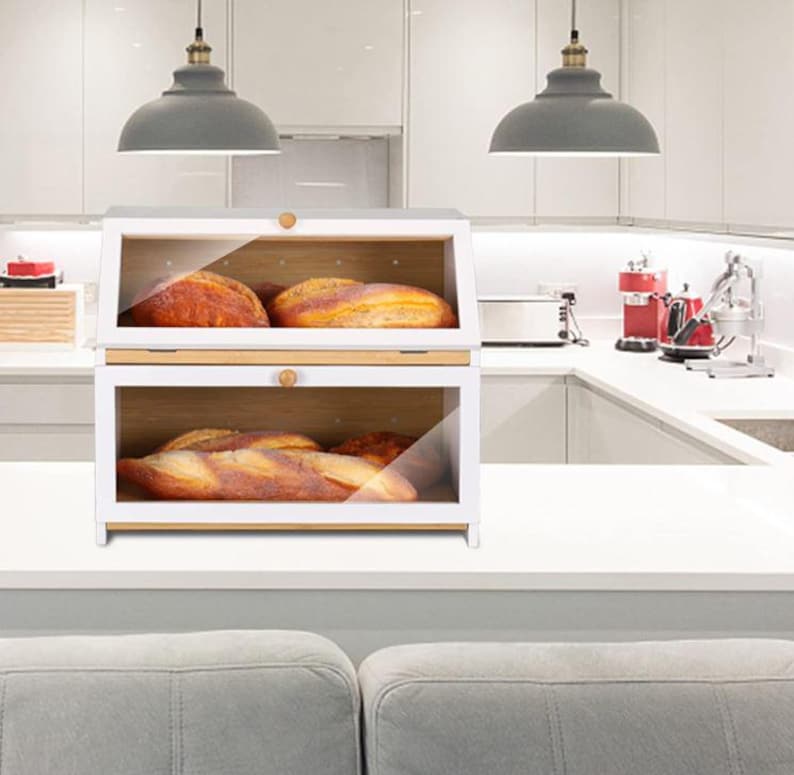 May include: A two-tiered white bread box with a clear glass front and a wooden interior. The box is filled with loaves of bread. The top of the box has a wooden handle. The bread box is on a white countertop in a kitchen.