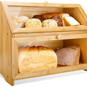 May include: A two-tiered bamboo bread box with clear glass doors. The top section holds muffins and sliced bread, while the bottom section contains a loaf of bread and sliced bread. The box has a natural wood finish and a simple, functional design.