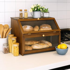 May include: A wooden bread box with two glass-fronted compartments, filled with various loaves of bread. The box sits on a white countertop, with jars of spices, pasta, and a bowl of lemons nearby. A small potted plant and flowers are on top.