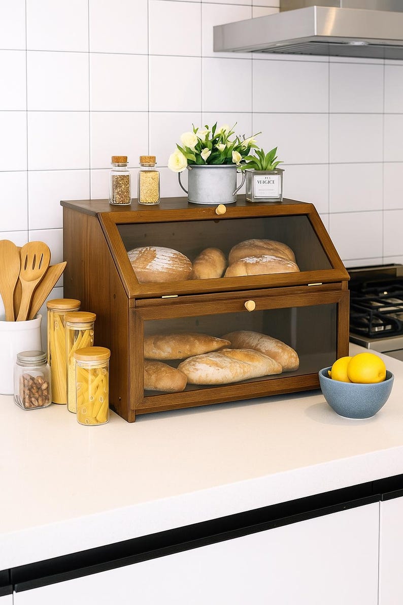 May include: A wooden bread box with two glass-fronted compartments, filled with various loaves of bread. The box sits on a white countertop, with jars of spices, pasta, and a bowl of lemons nearby. A small potted plant and flowers are on top.