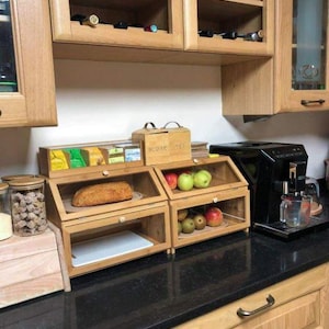 May include: Wooden kitchen storage units with glass doors, holding bread and fruit. Above, a cabinet with wine bottles. A coffee maker and a jar of cookies are also visible on the countertop. The cabinets are made of light-colored wood.