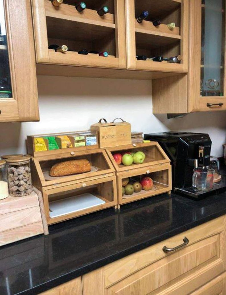 May include: Wooden kitchen storage units with glass doors, holding bread and fruit. Above, a cabinet with wine bottles. A coffee maker and a jar of cookies are also visible on the countertop. The cabinets are made of light-colored wood.