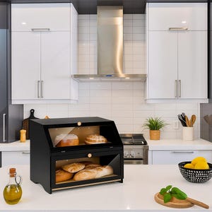 May include: A black wooden bread box with two transparent compartments, each holding loaves of bread. The box has a slanted top and wooden knobs. The kitchen setting includes white cabinets, a stainless steel range hood, and a countertop.