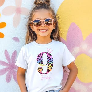 Könnte beinhalten: Ein weißes T-Shirt mit kurzen Ärmeln, das eine große Ziffer 9 mit buntem Leopardenmuster, eine kleine rosa Krone über der Ziffer und den Namen Emilia in rosa Schreibschrift unter der Ziffer zeigt.