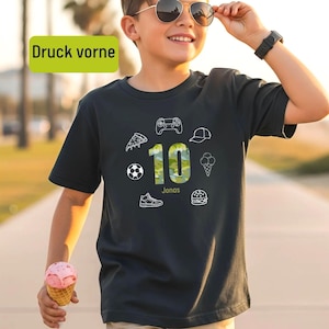 Könnte beinhalten: Ein dunkelgraues Kurzarm-T-Shirt mit der Zahl 10 in grünem Camouflage-Muster. Um die Zahl herum befinden sich weiße Strichzeichnungen von einem Gamecontroller, einer Baseballkappe, einem Eis, einem Burger, einem Sneaker, einem Fußball und einem Pizzastück.