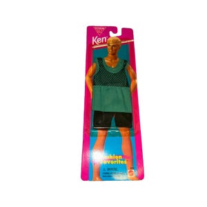 May include: A Ken doll in original packaging featuring a green mesh tank top and black shorts. The pink and blue card displays the text Ken and Fashion Favorites. The doll has blonde hair and tan skin, presented in a vertical plastic blister pack.