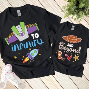 Puede incluir: Dos camisetas negras con gráficos coloridos. Una camiseta presenta un diseño de nave espacial con las palabras "To Infinity" y un cohete. La otra camiseta tiene un sombrero de vaquero, botas y las palabras "And Beyond".