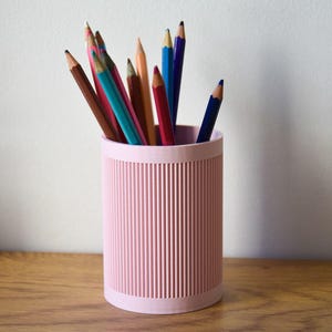 Puede incluir: Un portalápices rosa con detalles acanalados verticales, lleno de una variedad de lápices de colores. Los lápices son en tonos marrón, azul, rosa y naranja, creando una exhibición colorida. El portalápices está sobre una superficie de madera.