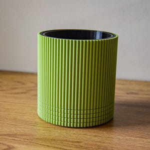 Peut inclure: Un pot de fleurs cylindrique vert citron avec des détails côtelés verticaux. L'intérieur est noir. Le pot est posé sur une surface en bois. Un design moderne et minimaliste pour la décoration intérieure.