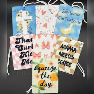 May include: A selection of air fresheners with diverse designs and text. One features a cross and floral elements, another has stethoscopes and hearts, and a third says "Silly Goose On The Loose" with a duck illustration. Other designs include text like "That Curl Hit Me" and "Mama Needs Coffee".