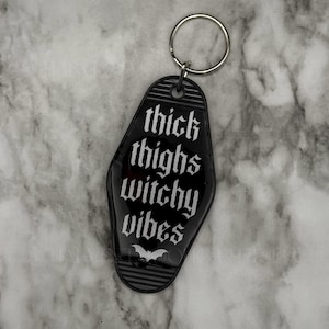 May include: A black motel-style keyring with a silver metal ring. The keyring features white text that reads thick thighs witchy vibes and a small white bat graphic at the bottom. The design has horizontal ribbed detailing at the top and bottom.