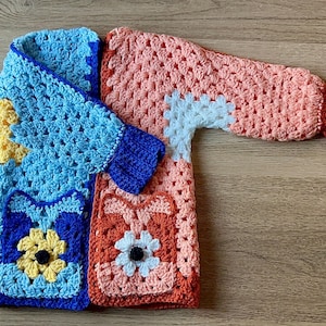 May include: A handmade crochet cardigan in a patchwork design. The cardigan features a mix of colors, including blue, peach, yellow, and white. The design includes floral motifs and a textured stitch pattern. The garment is laid flat on a wooden surface.