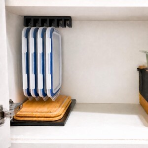 May include: A black plastic under-shelf storage rack mounted to the top of a cabinet, holding four rectangular food storage container lids with white frames and blue silicone seals, positioned above two stacked rectangular bamboo cutting boards.