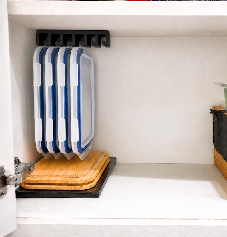May include: A black plastic under-shelf storage rack mounted to the top of a cabinet, holding four rectangular food storage container lids with white frames and blue silicone seals, positioned above two stacked rectangular bamboo cutting boards.