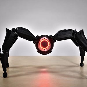 May include: A black, mechanical spider-like robot with six articulated legs and a central body. The robot has a glowing red eye and is positioned on a light-colored surface. The design suggests a futuristic or sci-fi theme.