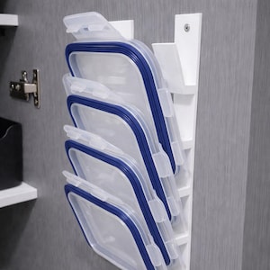 May include: A white plastic organiser holds several clear food storage containers with blue trim. The containers are stacked vertically, ready for use. The organiser is mounted on a wall inside a cabinet.