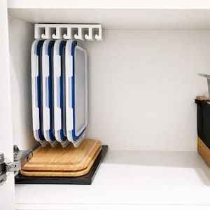 May include: A white plastic under-shelf storage rack with six slots holds four rectangular food container lids with white frames and blue silicone seals. Below the rack, three stacked wooden cutting boards rest on a black base.