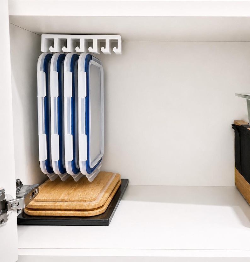 May include: A white plastic under-shelf storage rack with six slots holds four rectangular food container lids with white frames and blue silicone seals. Below the rack, three stacked wooden cutting boards rest on a black base.