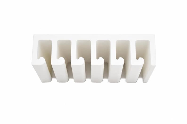 May include: A white wall-mounted organizer featuring six evenly spaced vertical slots with curved hook-like ends. The structure is rectangular with a solid top bar and uniform open channels designed for holding items in a linear arrangement.